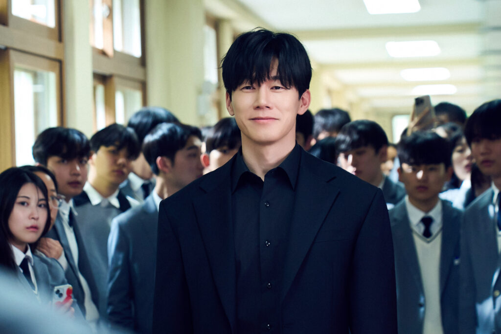 "A gentleman in an all-black suit smiles with ease while a group of students look at him with anticipation."